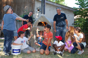 Ein Mitarbeiter der Firma ORTNER und eine Gruppe Kinder steht vor einem beheizten ORTNER Holzbackofen.