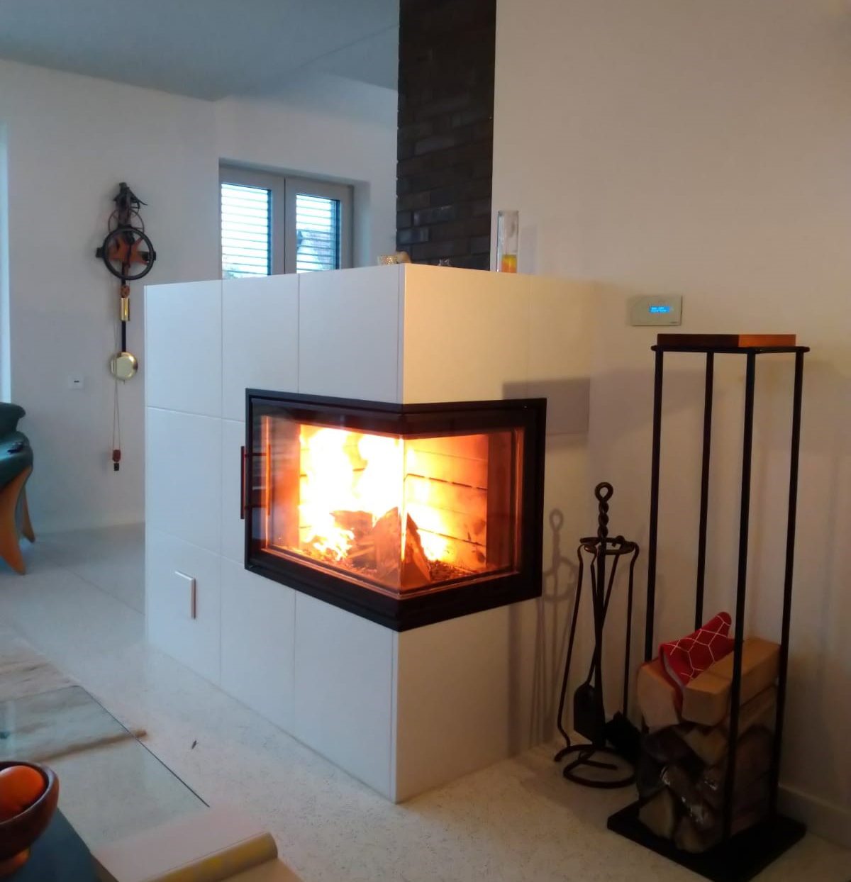 Ein Speicherofen mit weißer Keramik und in dem ein Feuer brennt. Ein Feuerbesteck steht davor.